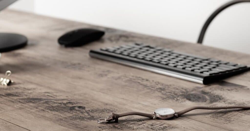Holzschreibtisch mit Computertastatur, Maus, Büroklammer aus Metall und einer Armbanduhr mit braunem Armband.