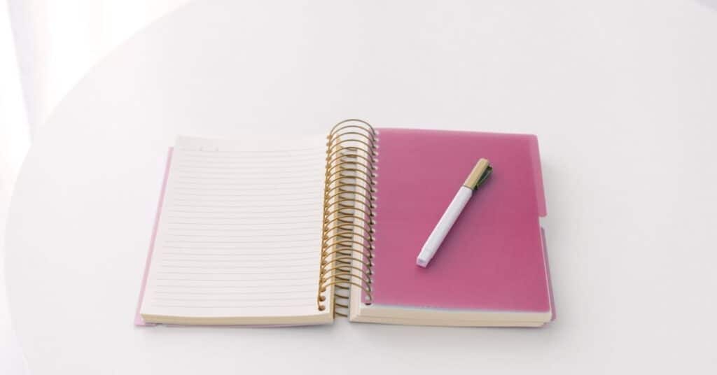 Aufgeschlagenes Spiralnotizbuch mit rosa Einband und einem darauf liegenden Stift, auf einer weißen Fläche platziert.