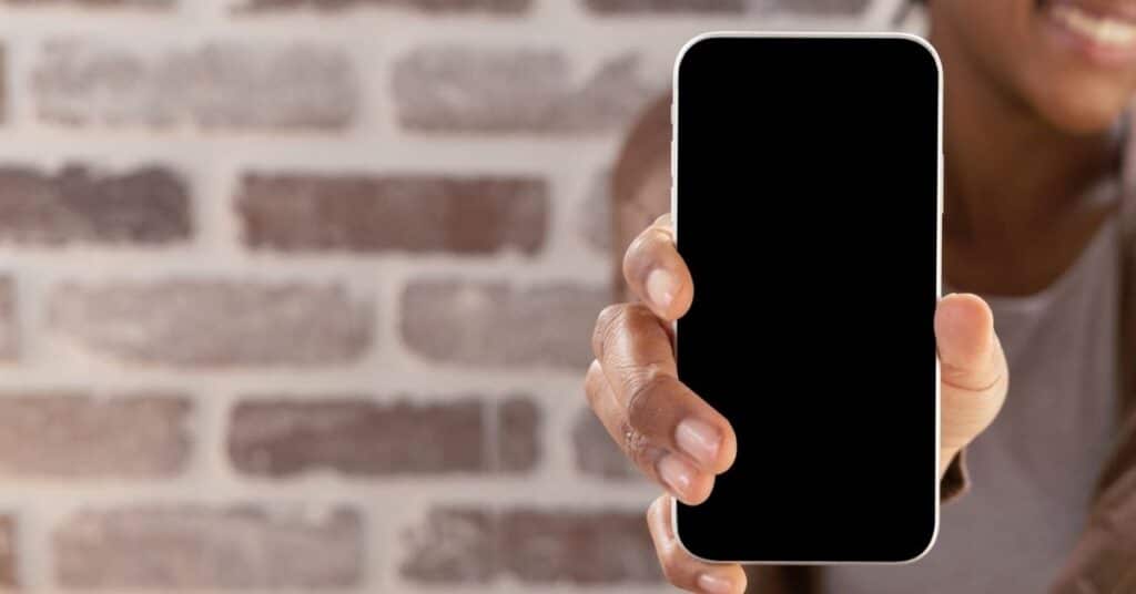 Person, die ein Smartphone mit schwarzem Bildschirm vor einem Ziegelwandhintergrund hält.