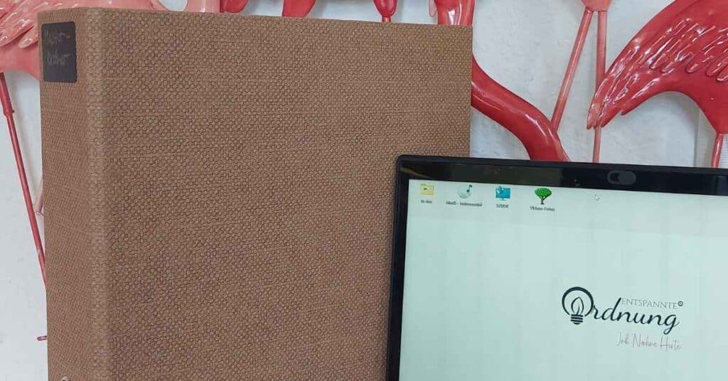 Ein Laptop mit dem Wort „Ordnung“ neben einem großen, strukturierten braunen Buch mit einem Etikett aus Tafelfolie. Im Hintergrund sind rosa Flamingo-Dekorationen zu sehen.