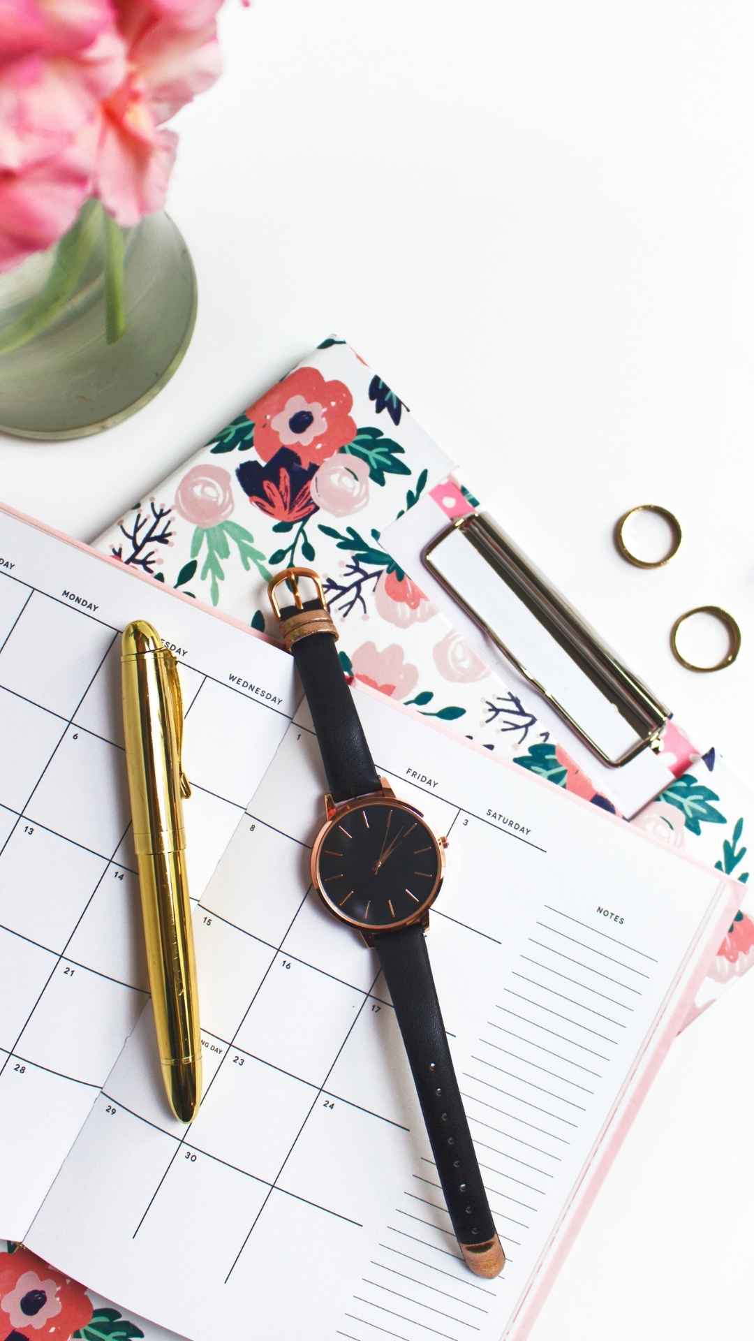 Ein goldener Stift, eine schwarze Armbanduhr und goldene Ringe auf einem offenen Blumenplaner. Rosa Blumen stehen in einer Vase daneben.